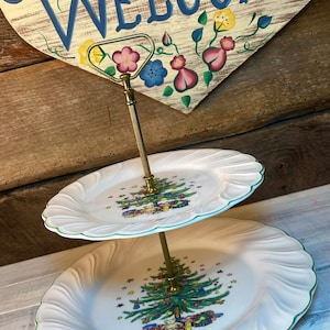 May include: Two-tiered white ceramic serving stand with a gold metal stand. Each tier features a green border and a Christmas tree design with presents and a teddy bear.