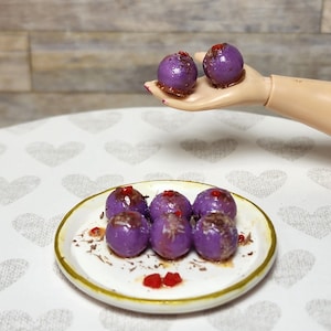 May include: A miniature plate with a gold rim holds six purple, round food items topped with red accents. Two similar items are held in a doll's hand. The food items are on a white surface with a heart pattern.
