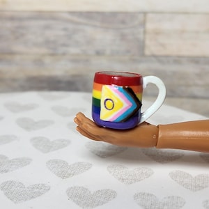 May include: A small, rainbow-colored mug with a white handle. The mug features a Progress Pride flag design. The mug is held in a brown hand, against a backdrop of a light-colored, heart-patterned surface and a wooden wall.
