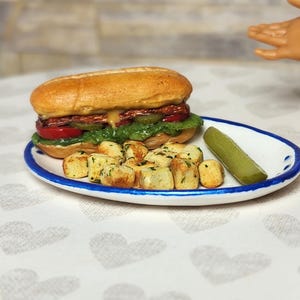 May include: Miniature sandwich with lettuce, tomato, pickles, and cheese on a bun, served with fried potato cubes and a pickle spear on a white plate with a blue rim. The food is arranged on a heart-patterned tablecloth.