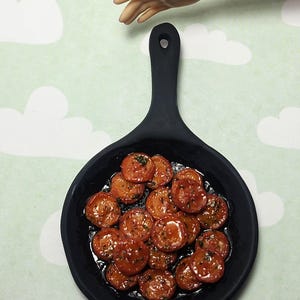 May include: Miniature black frying pan filled with cooked, sliced sausage. The sausage is a reddish-brown color and appears to be seasoned with herbs. The background is a light green color with white cloud shapes.
