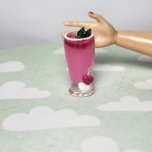 May include: A miniature pink drink in a clear glass, topped with a dark red layer and green leaves. Two heart-shaped decorations, one red and one white, adorn the base. The drink is set against a light green background with white cloud patterns.