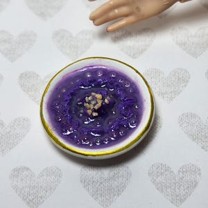 May include: Miniature ceramic bowl filled with a vibrant purple substance, possibly food, with a gold rim. The bowl's contents have a textured appearance, and a few small, light-colored pieces are visible in the center. The bowl is on a white surface with heart-shaped patterns.