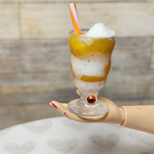 May include: A layered smoothie in a clear glass with a decorative stem. The smoothie has layers of yellow, white, and speckled colors, topped with a dollop of white cream and a striped straw. The glass is held by a doll hand.