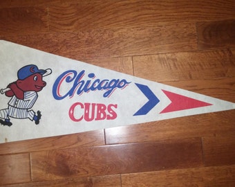May include: Vintage Chicago Cubs pennant featuring a cartoon bear wearing a baseball cap and holding a bat. The pennant is white with blue and red accents and the text "Chicago Cubs".