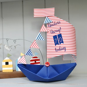 May include: A blue paper sailboat with red and white striped sails. The sails have the text "Anchors Away! 70 today" printed on them. The sailboat is decorated with a red button and a string of red and blue triangular flags.