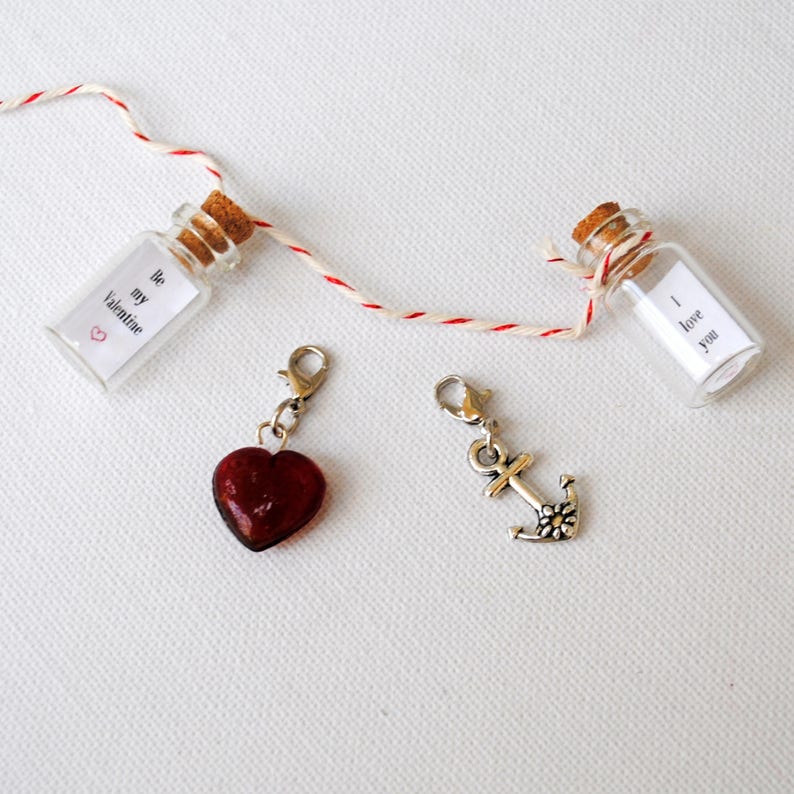 May include: Two small glass bottles with cork stoppers, each containing a rolled up piece of paper with a message. The messages read "Be my Valentine" and "I love you".  A red heart charm and a silver anchor charm are also pictured.