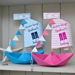 May include: Two paper sailboats decorated for a 50th birthday party. The blue sailboat has a white and blue striped sail with the text "Anchors Away! Andrew's 50 today". The pink sailboat has a pink and white striped sail with the text "Anchors Away! Andrew's 50 today".
