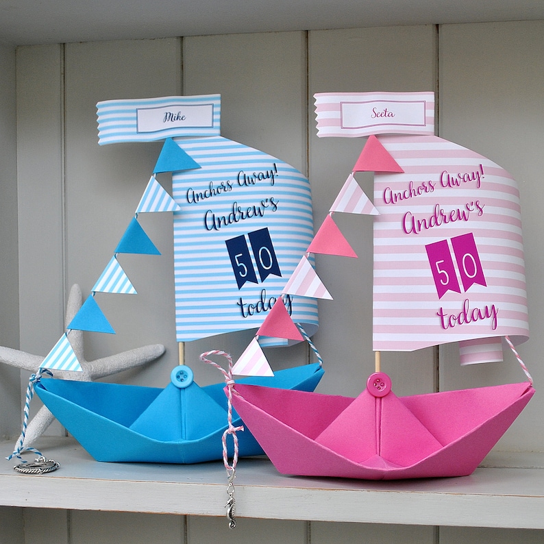 May include: Two paper sailboats decorated for a 50th birthday party. The blue sailboat has a white and blue striped sail with the text "Anchors Away! Andrew's 50 today". The pink sailboat has a pink and white striped sail with the text "Anchors Away! Andrew's 50 today".