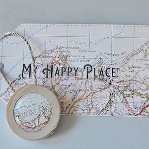 May include: A tag-shaped paper item with a map design and the words "MY HAPPY PLACE!" printed on it. A silver-colored magnifying glass is attached to the tag. The map shows roads, bodies of water, and place names.