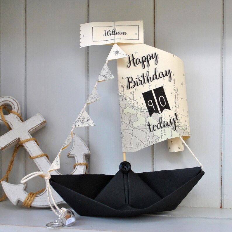 May include: A black paper sailboat with a white map sail that says "Happy Birthday 90 today!" and a small white flag that says "William".