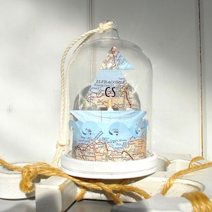 May include: A glass dome with a white rope handle contains a paper sailboat made from a vintage map of Ilfracombe, England. The sailboat is surrounded by blue paper waves and sits on a white base.