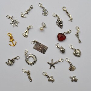 May include: A collection of 19 silver charm pendants with various designs, including anchors, seashells, a cupcake, a heart, a starfish, a turtle, a seahorse, a life preserver, a postcard, and a teacup. The charms are on a white background.