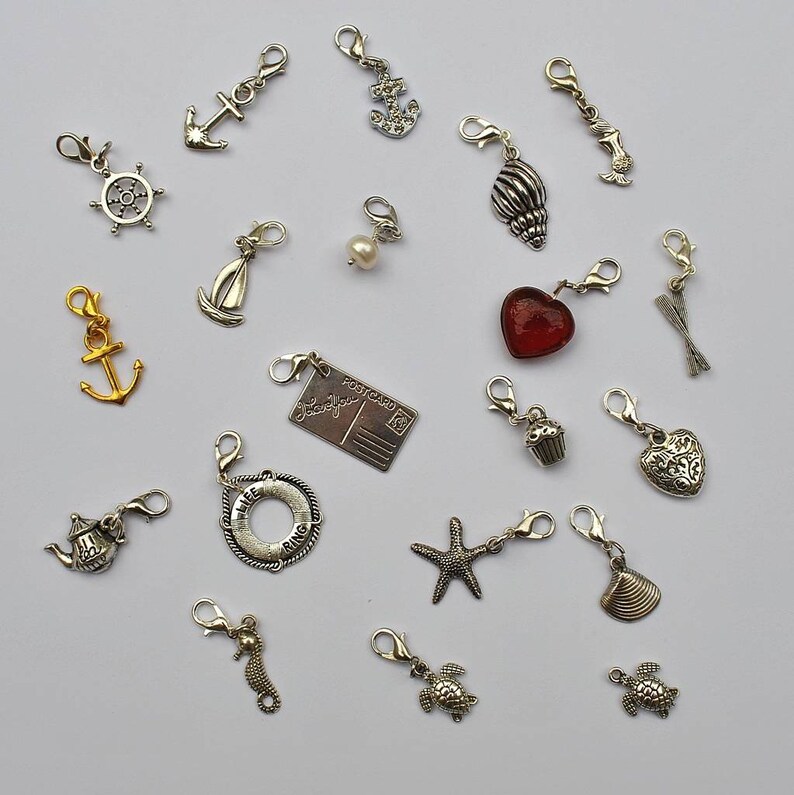 May include: A collection of 19 silver charm pendants with various designs, including anchors, seashells, a cupcake, a heart, a starfish, a turtle, a seahorse, a life preserver, a postcard, and a teacup. The charms are on a white background.