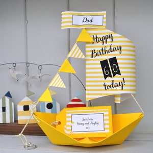 May include: A yellow paper sailboat with a white and yellow striped sail. The sail has the text "Happy Birthday 60 today!" and a yellow tag with the text "Love from Krissy and Hayley xxxx".