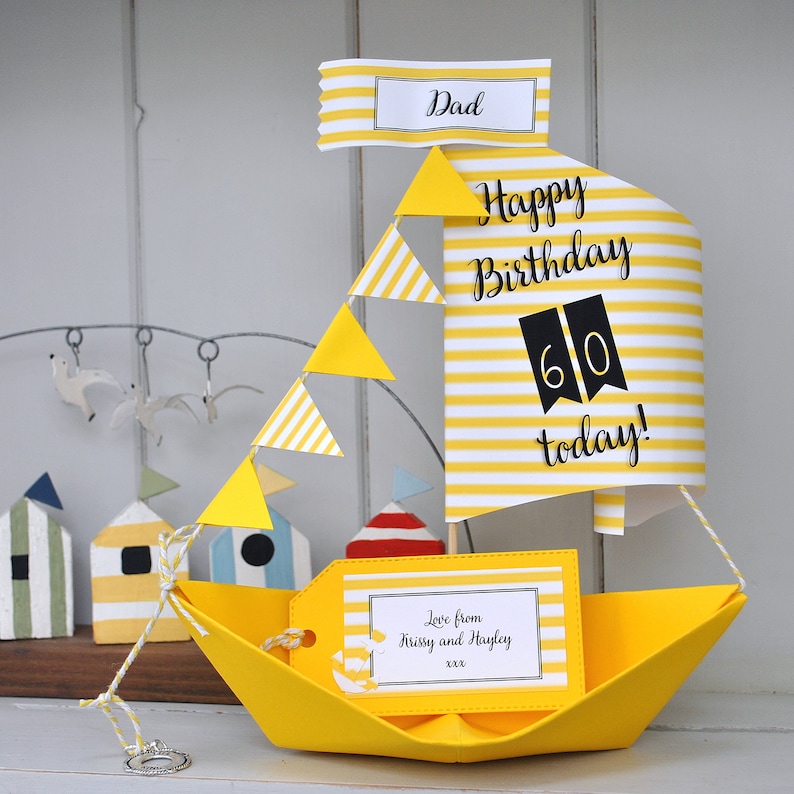 May include: A yellow paper sailboat with a white and yellow striped sail. The sail has the text "Happy Birthday 60 today!" and a yellow tag with the text "Love from Krissy and Hayley xxxx".