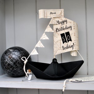 May include: A paper ship birthday decoration with a map sail, a black paper boat, and a globe. The sail says "Happy Birthday 50 today!" and has a tag that says "Mark".