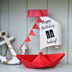 May include: A red paper sailboat with a white sail that says "Happy Birthday 58 today!" The sailboat has a red ribbon and red paper flags. There is a white wooden anchor in the background.