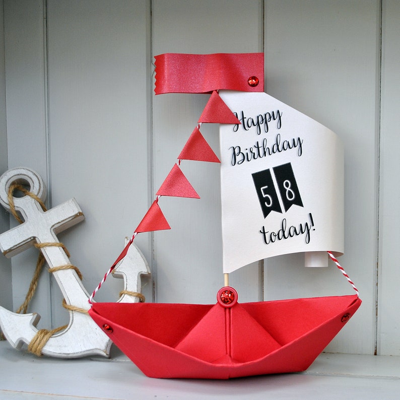 May include: A red paper sailboat with a white sail that says "Happy Birthday 58 today!" The sailboat has a red ribbon and red paper flags. There is a white wooden anchor in the background.