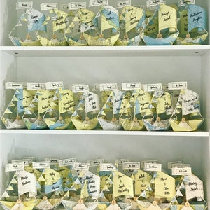 May include: A shelf with three rows of small paper sailboats, each with a name card attached. The sailboats are made of maps and are decorated with buttons. The names on the cards are for a wedding reception.