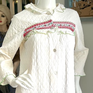 Pode incluir: Um roupão acolchoado branco vintage com um padrão de losangos, exibido em um manequim. O roupão apresenta um painel decorativo com bordados vermelhos e verdes, uma gola com babados e fechos de botões. As mangas e a gola são aparadas em verde.