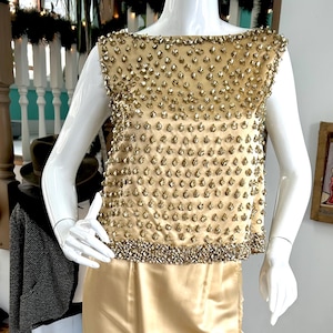 May include: A gold-colored, two-piece formal dress on a mannequin. The top is sleeveless and heavily embellished with small, gold-colored beads. The skirt is a long, straight, satin-like material with a side slit.