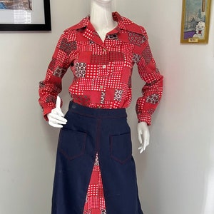 May include: A vintage two-piece outfit displayed on a mannequin. The red shirt features a white and black geometric pattern. The navy blue skirt has red stitching and a split front, showing a matching red patterned fabric.