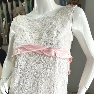 Puede incluir: Vestido blanco sin mangas de encaje con cinturón de cinta de raso rosa. El vestido se exhibe en un maniquí. El encaje tiene un patrón detallado y calado. La cinta rosa añade un toque de color.