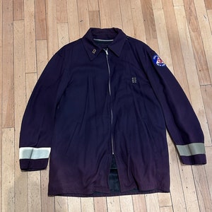 Puede incluir: Chaqueta morada oscura con cremallera completa, rayas reflectantes en las mangas y cuello. La chaqueta tiene un parche en la parte superior del brazo con texto y un logotipo. La chaqueta está tendida sobre una superficie de madera.