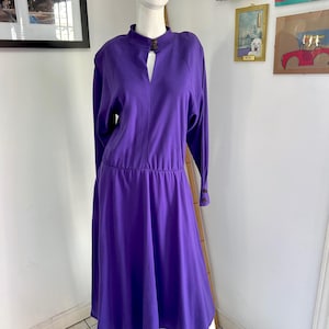 May include: A long-sleeved, knee-length purple dress with a high collar and a keyhole neckline. The dress has a fitted bodice and a flared skirt. The sleeves have button details at the cuffs. The dress is displayed on a mannequin.