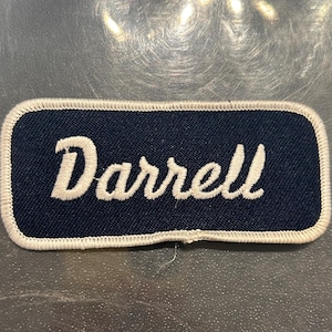 May include: A white embroidered name tag with a navy blue background. The name "Darrell" is stitched in white.