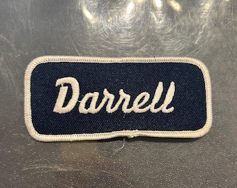 May include: A white embroidered name tag with a navy blue background. The name "Darrell" is stitched in white.