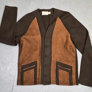May include: A brown cardigan with a V-neck and long sleeves. The front features brown suede panels and pockets, with a button-up closure. The sleeves and back are a darker brown knit material. A small tag is visible near the neckline.