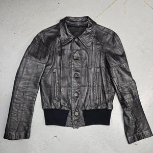 May include: Black leather jacket with a quilted design. The jacket features a button-up front, a collar, and a black ribbed hem and cuffs. The jacket has a classic bomber style.