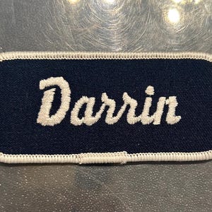 May include: A navy blue embroidered name tag with white stitching around the edges. The name "Darrin" is embroidered in white on the tag.