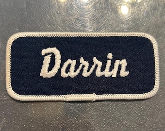 May include: A navy blue embroidered name tag with white stitching around the edges. The name "Darrin" is embroidered in white on the tag.