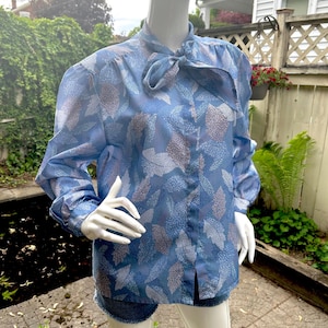May include: Light blue blouse with a leaf pattern. The long-sleeved top features a bow at the neck and a button-down front. The blouse is displayed on a mannequin.