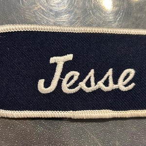 May include: A navy blue fabric patch with white stitching around the edges. The word "Jesse" is embroidered in white on the patch.