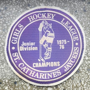 May include: Purple and white embroidered patch with the text "Girls Hockey League St. Catharines Jaycees" and "Junior Division Champions 1975-76". A girl hockey player is depicted in the center.