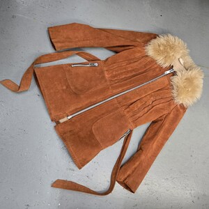 May include: A brown suede jacket with a faux fur collar. The jacket features a zipper closure, two front pockets, and a matching belt. The sleeves are long, and the jacket has a fitted waist. The jacket is laid flat on a gray surface.