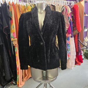 May include: A black textured jacket with a shawl collar and a single button fastening. The jacket has long sleeves and a fitted waist. The jacket is displayed on a mannequin in a vintage clothing shop.
