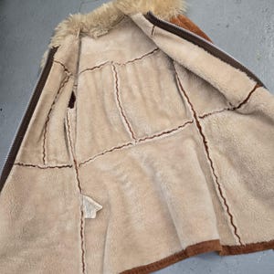 May include: Open view of a brown and tan shearling jacket with a faux fur collar. The jacket features a zipper closure and visible stitching details. The interior is lined with a soft, plush material.