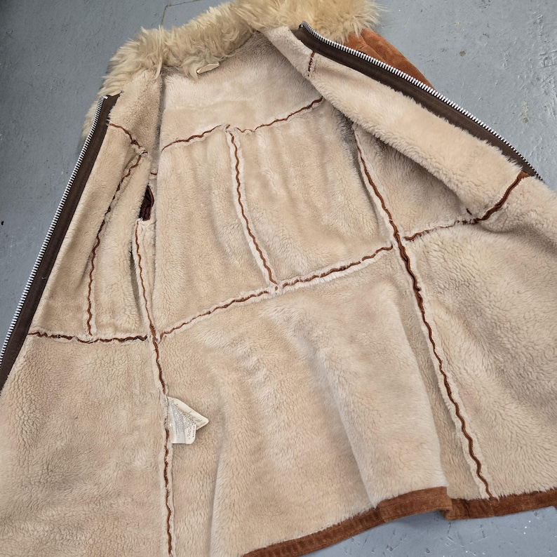 May include: Open view of a brown and tan shearling jacket with a faux fur collar. The jacket features a zipper closure and visible stitching details. The interior is lined with a soft, plush material.