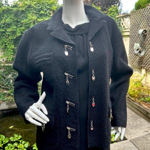 May include: A black wool jacket with silver zip and charm details. The jacket features a classic collar, a single pocket, and decorative charms. The jacket is displayed on a mannequin.