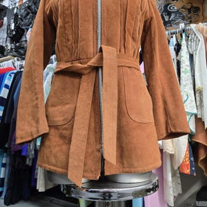 May include: A brown suede jacket with a zip-up front, long sleeves, and a matching belt. The jacket has two front pockets and is displayed on a mannequin. The jacket is a vintage style.