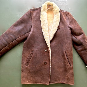 May include: A brown sheepskin coat with a shearling lining and a single button fastening. The coat has two pockets on the front and a collar that can be worn up or down.