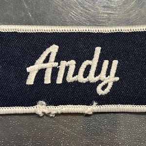 May include: A navy blue fabric name tag with white stitching around the edges. The name "Andy" is embroidered in white cursive lettering.