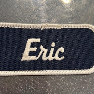 May include: A navy blue name tag with white stitching around the edges. The name "Eric" is embroidered in white on the tag.