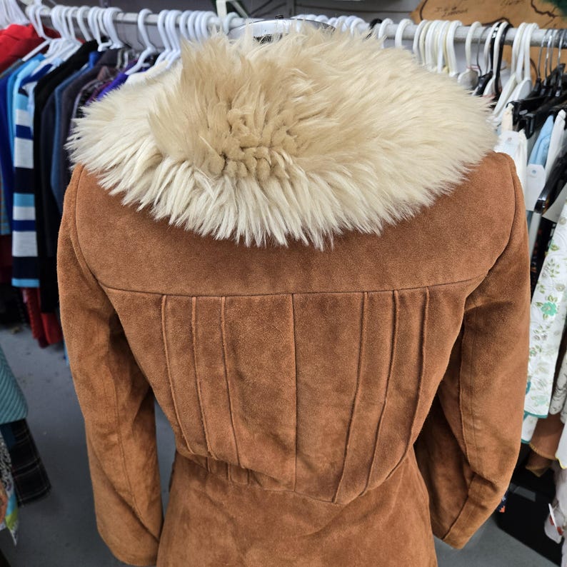 May include: A brown suede jacket with a cream-colored faux fur collar. The back of the jacket features vertical pleats and a cinched waist. The jacket is displayed on a rack with other clothing.