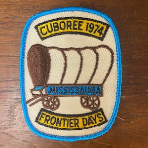 May include: A blue and yellow embroidered patch with a covered wagon and the text "Cuboree 1974", "Mississauga", and "Frontier Days".